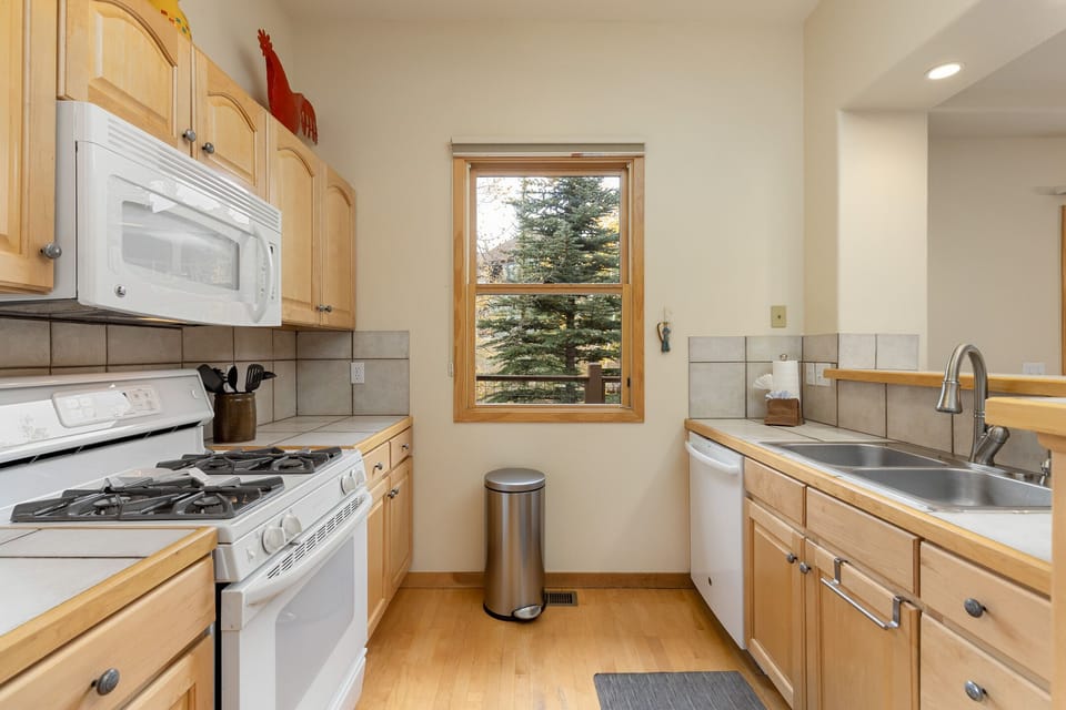 A kitchen with wooden cabinets, a white gas stove, white microwave, sink, dishwasher, trash bin, and a window with a view of trees outside.