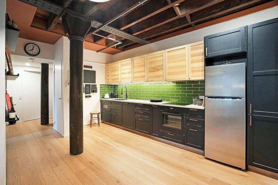Timber beams enclose the galley kitchen with sleek, green subway tiles, dark cabinetry and electric appliances.