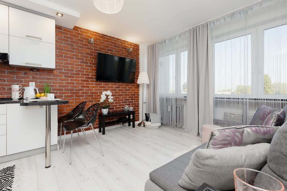 An open-plan kitchen and living area with a grey sofa, dining table, and large windows letting in natural light.
