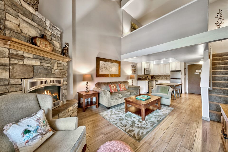 Relax in your open-concept living space where warm wood floors meet cozy seating and a crackling stone fireplace creates the perfect mountain retreat atmosphere.
