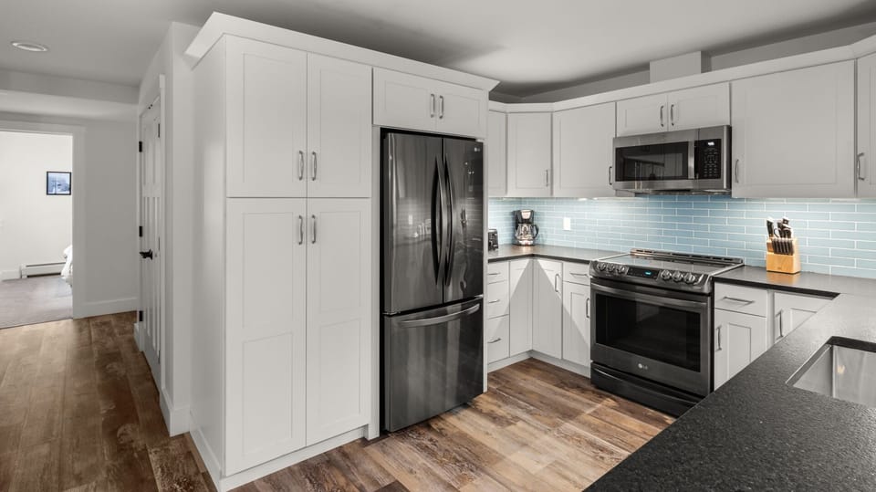 Modern kitchen featuring white cabinetry, stainless steel appliances, a blue tile backsplash, and a wooden floor. The room is well-lit, with a corridor visible leading to another part of the home.