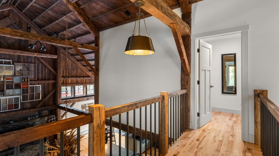 A rustic loft with a wooden railing, exposed beams, a metal hanging light fixture, and access to a door on the right. The area overlooks a lower level with a combination of windows and wood paneling.