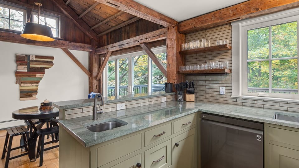 A modern kitchen with green cabinetry, granite countertops, and stainless steel appliances. Wooden beams accent the ceiling and walls. A dining area with a small round table and chairs is in the background.