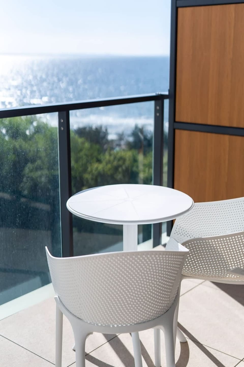 A cozy dining area on the balcony with chairs and a table, set against a backdrop of greenery and the coastline.
