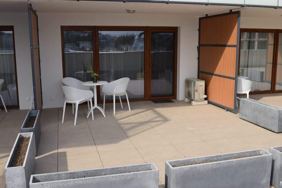 A spacious terrace with a tiled floor, modern outdoor furniture including a round table and white chairs, and planters adding greenery to the area.
