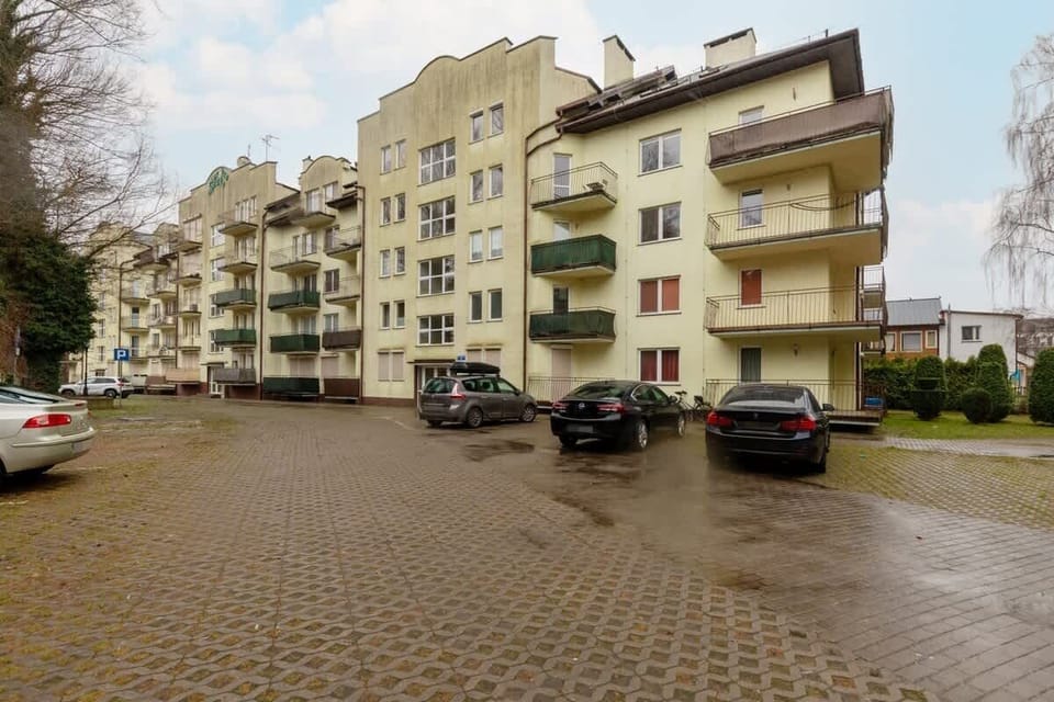 An exterior view of a residential building with a paved parking area and greenery around the entrance.
