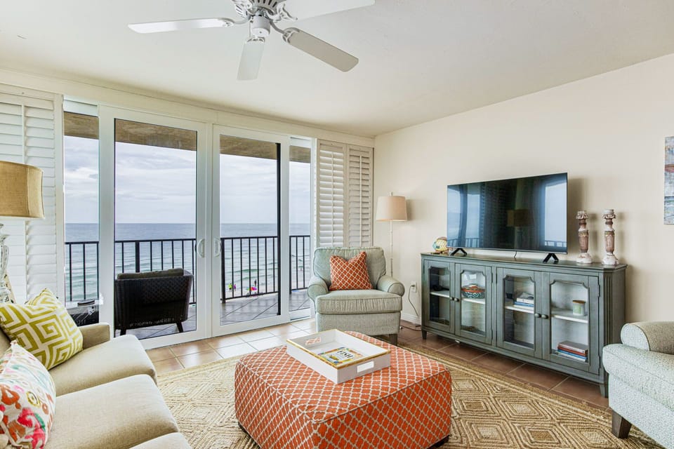 he living room at Lei Lani 401, Orange Beach — flat-screen TV, ceiling fan, and panoramic Gulf views framed by large sliding glass doors connecting to the wraparound balcony.