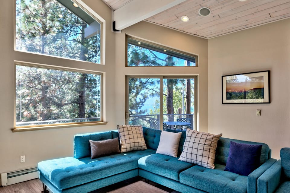 Teal sectional with patterned pillows faces sliding glass doors leading to a balcony framed by tall pine trees.