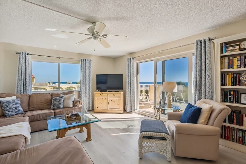 - Bright living room with ocean view windows bringing in natural sunlight daily  
- Comfortable seating with sofa chairs TV and bookshelf for relaxing moments  
- Open layout makes it perfect for families to unwind after beach adventures  
