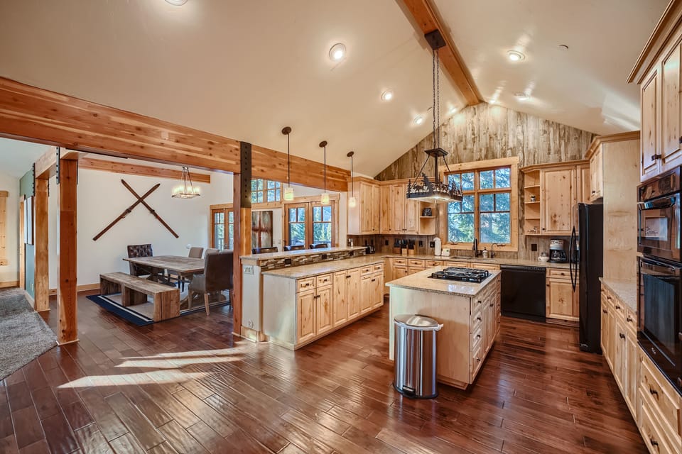 Spacious kitchen and dining area with wooden cabinetry, hardwood floors, an island with a stove, large windows, and a rustic dining table. Wall-mounted skis decorate the dining space.