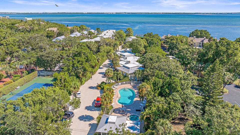 Aerial view of a residential area with a central swimming pool, tennis court, lush greenery, and adjacent water body under a clear sky.