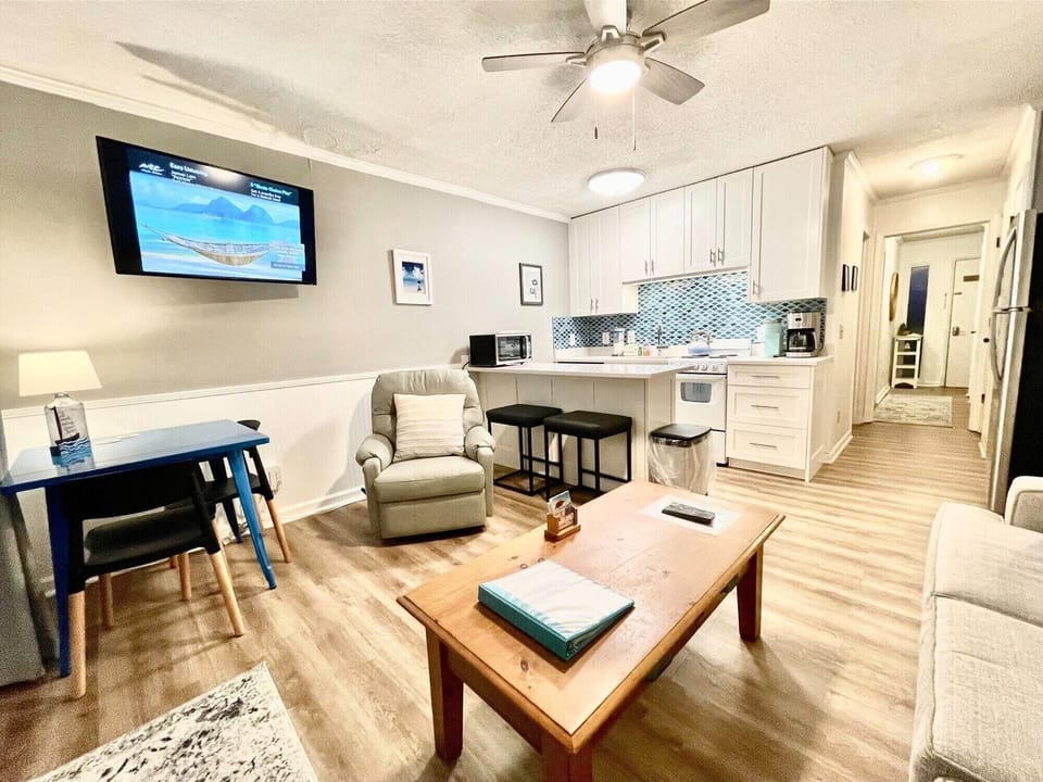 A cozy living room and kitchen area with a TV, armchair, and wooden coffee table. The kitchen has white cabinets, a small counter with stools, and various appliances.