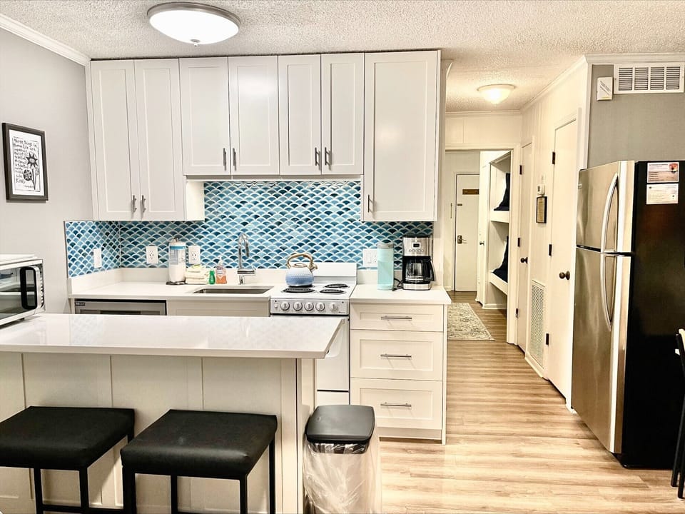 Modern kitchen with white cabinetry, blue tiled backsplash, stainless steel appliances, and a central island with two black stools. Adjacent hallway and wall art visible.