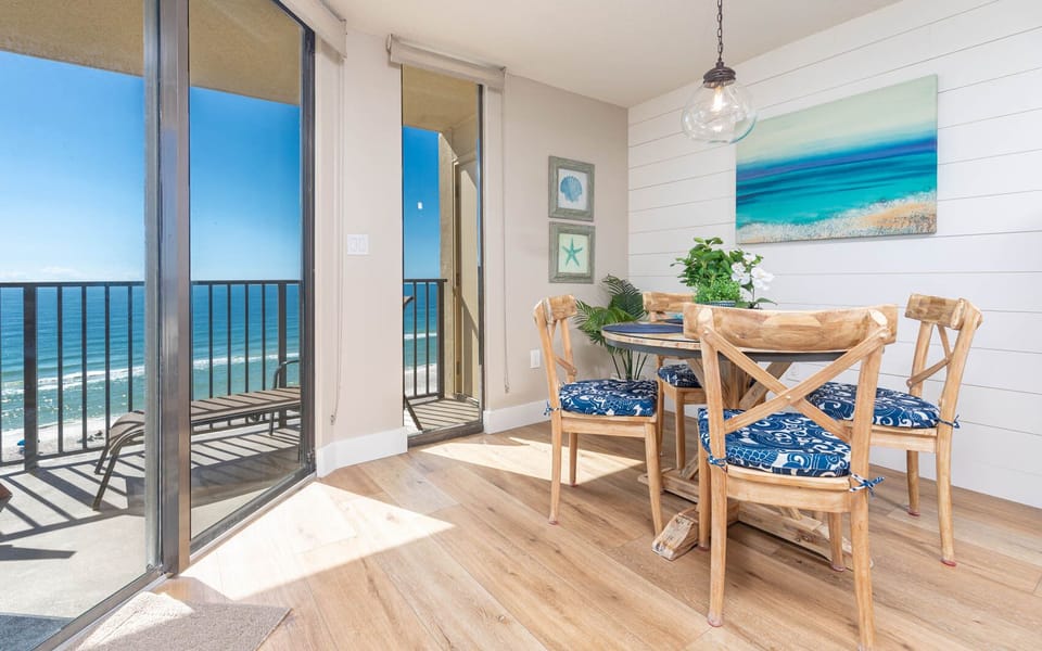 Bright dining area features a round table with blue-patterned chairs, offering a stunning view of the beach from the balcony