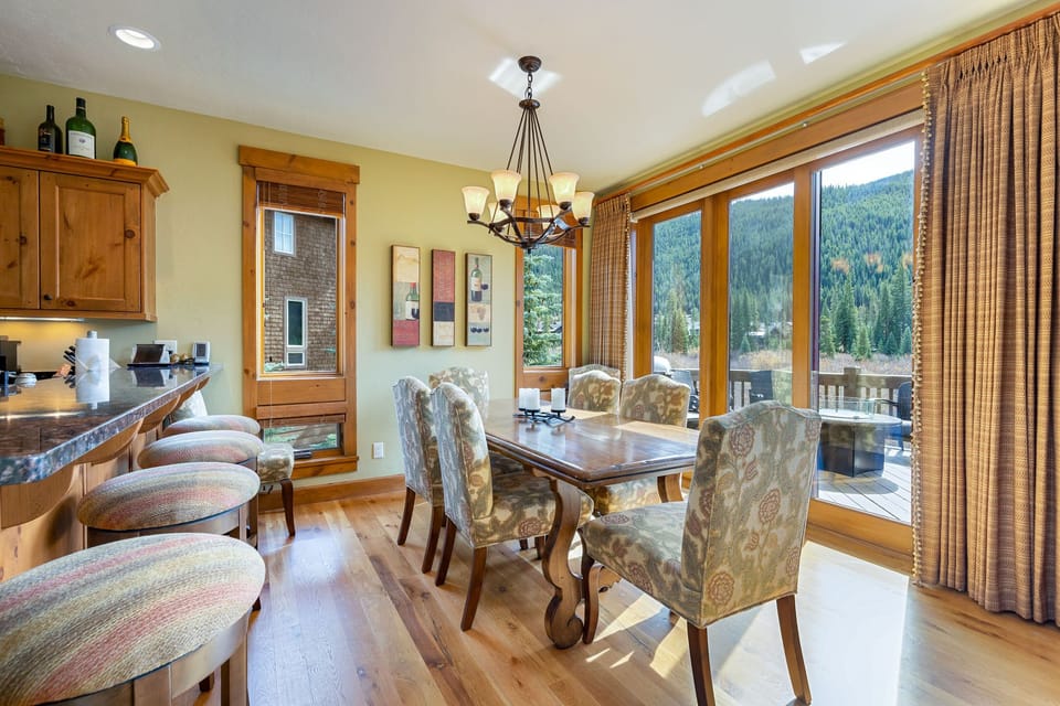 A dining room with a wooden table, six upholstered chairs, and a chandelier. A breakfast bar with stools is visible to the left. Large glass doors provide a view of a deck and wooded hillside outside.